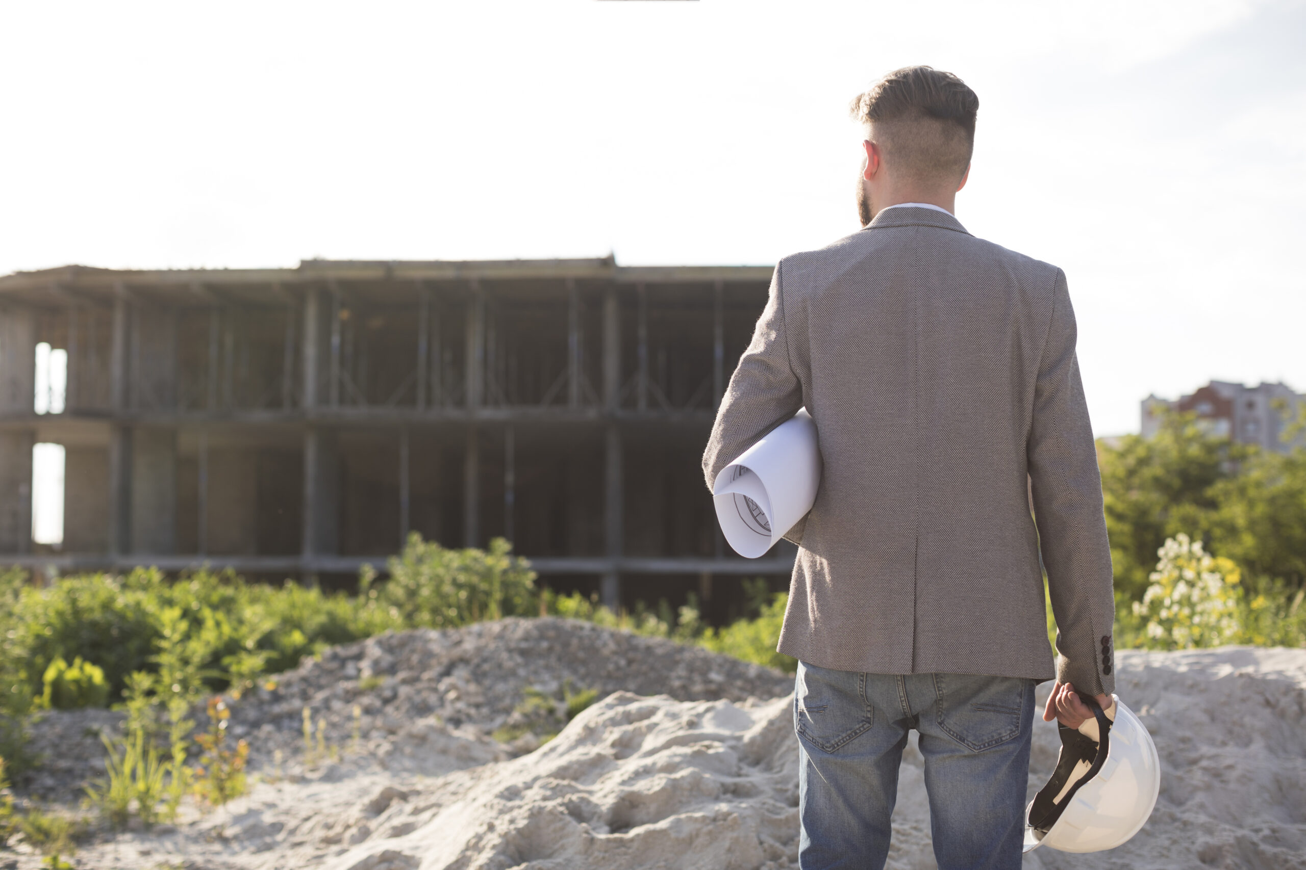 rear-view-male-architecture-holding-blueprint-hard-hat-looking-construction-site
